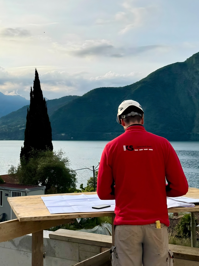 Un dipentende dell'impresa edile con maglione rosso con il logo dell'azienda studia i piani di costruzione con vista sul lago di Como