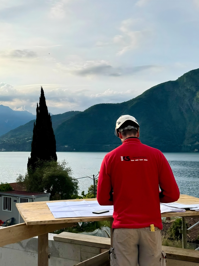 Ein Mitarbeiter der Baufirma in rotem Pullover mit Firmenlogo studiert Baupläne mit Bilck auf den Como-See. 
